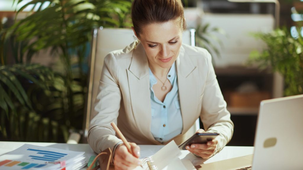 Donna che lavora alla scrivania prendendo appunti e consultando il telefono e si prepara per una transizione professionale graduale e consapevole